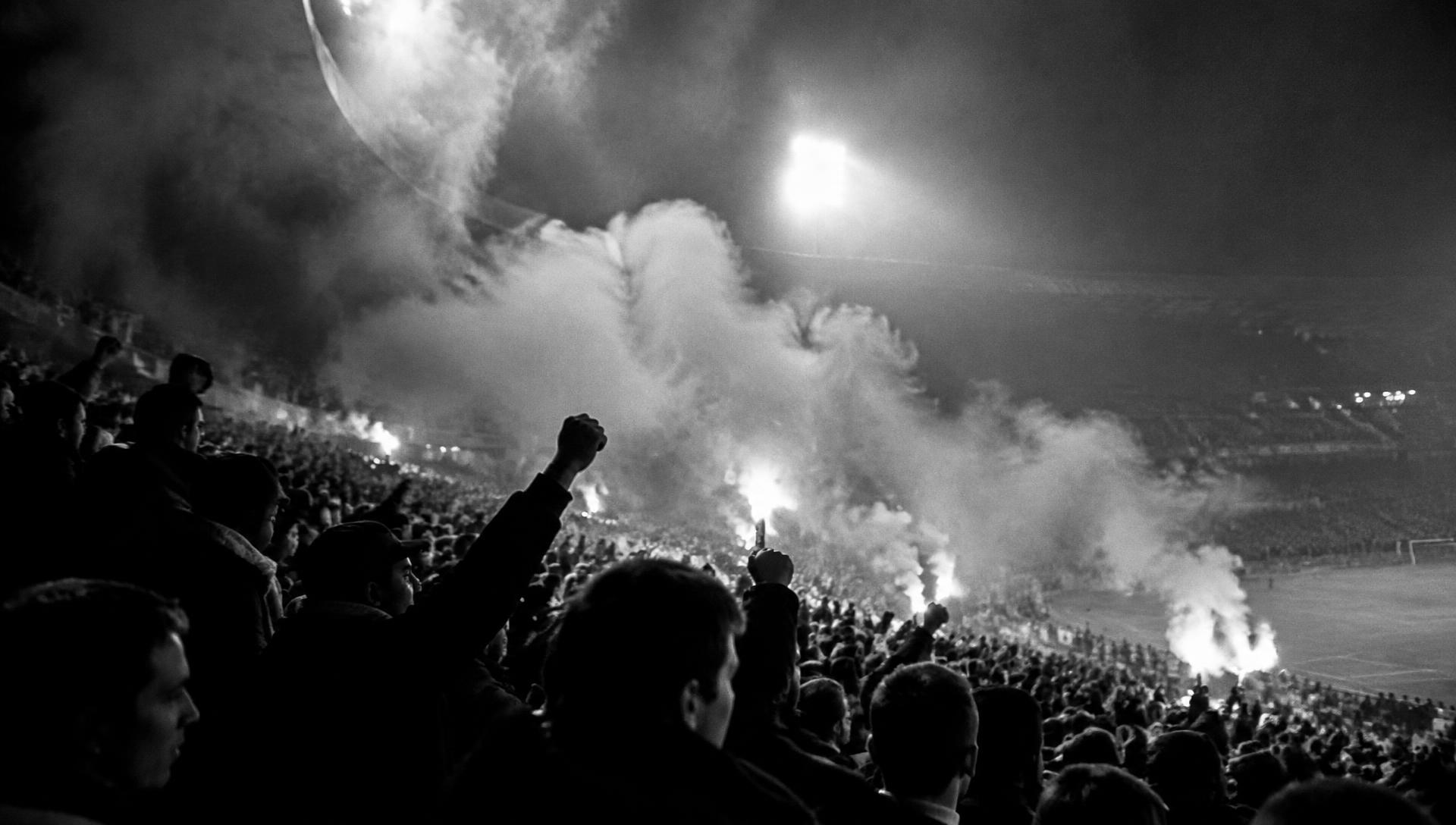 Supporters with raised fists and flares on a stadium terrace at night